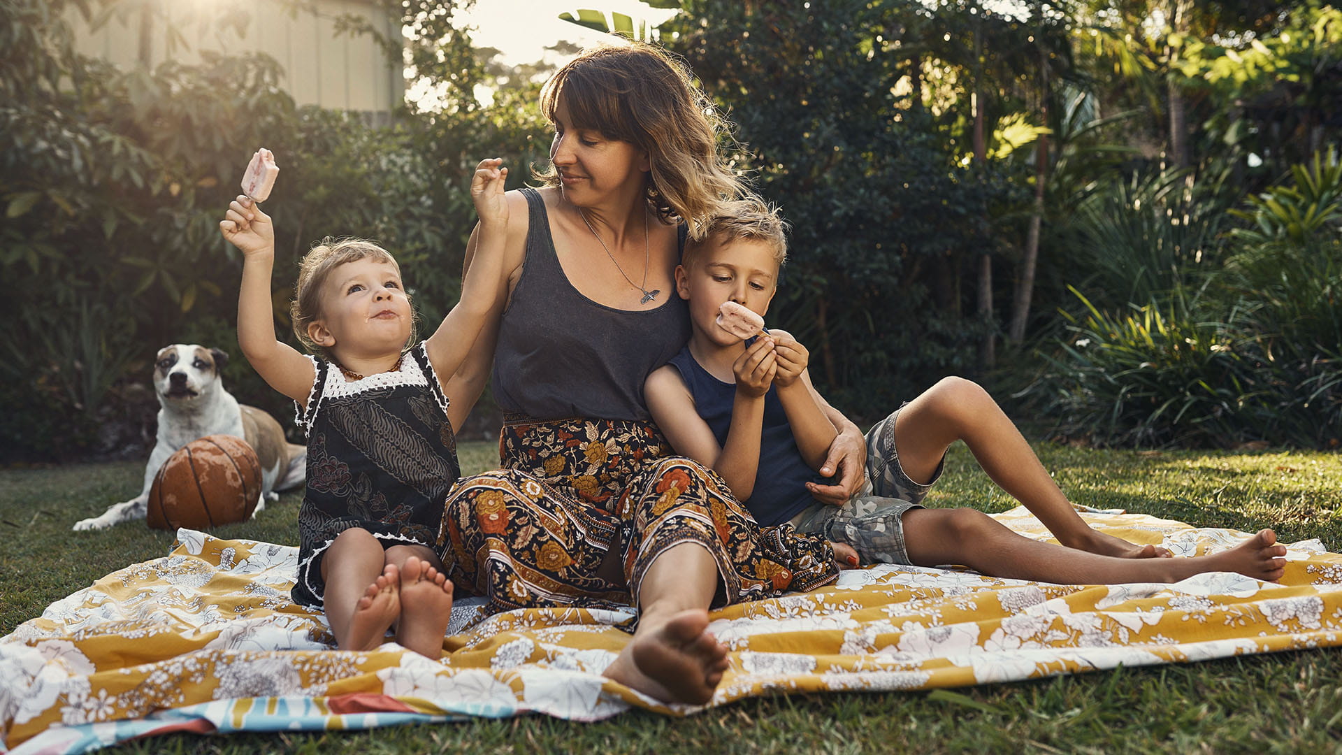A woman shares a blanket with two children in a sunny backyward while they all eat ice-cream. A dog sits in the background.