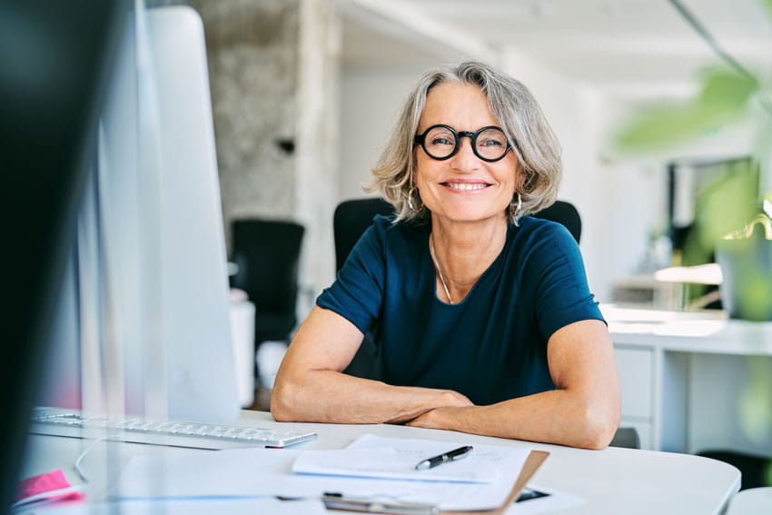 Portrait of confident smiling mature businesswoman with glasses sitting at a desk.