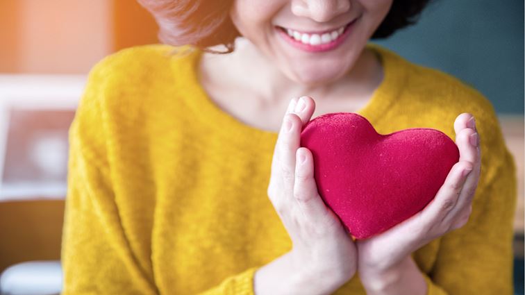 A woman wearing a yellow jumper is holding a plush red heart and smiling